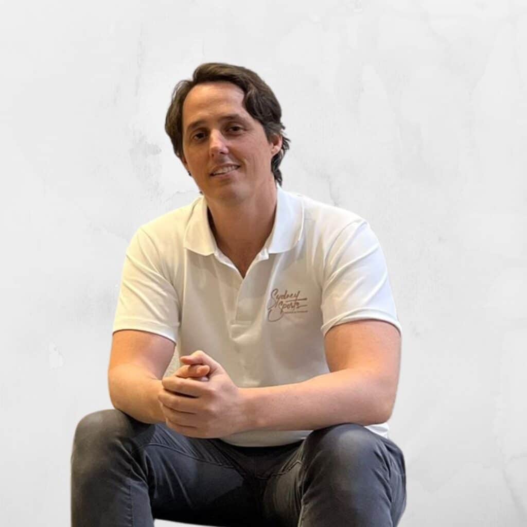 A person sits wide-legged wearing a white polo shirt with "Sydney Sports" embroidered, set against a plain, light gray background.