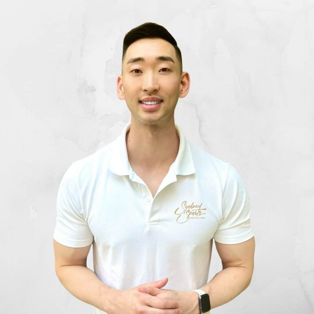 A person wearing a white polo shirt with "Sydney Sports & Exercise" text stands smiling against a plain white background, with hands gently clasped.