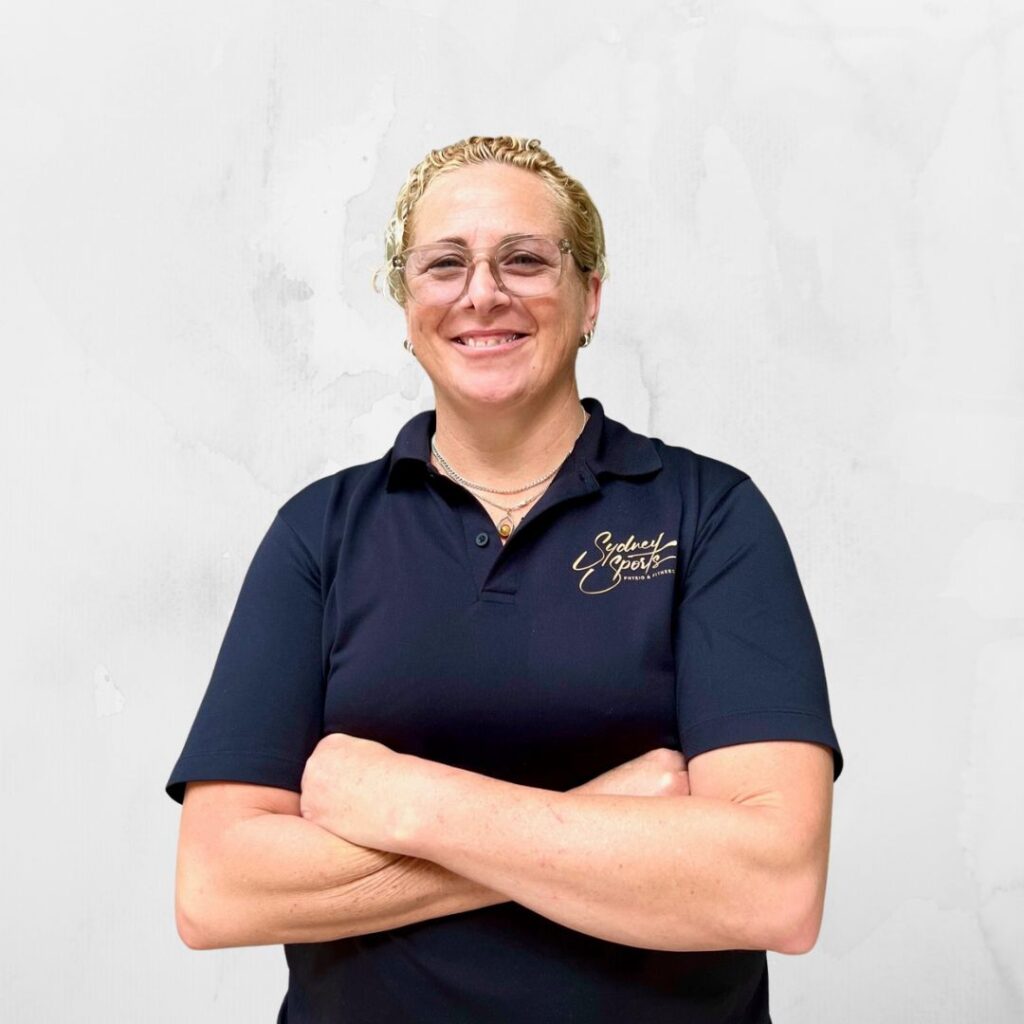 A person with short blond hair, wearing glasses and a dark shirt with "Sydney Sports" logo, stands smiling with arms crossed against a plain white textured wall.