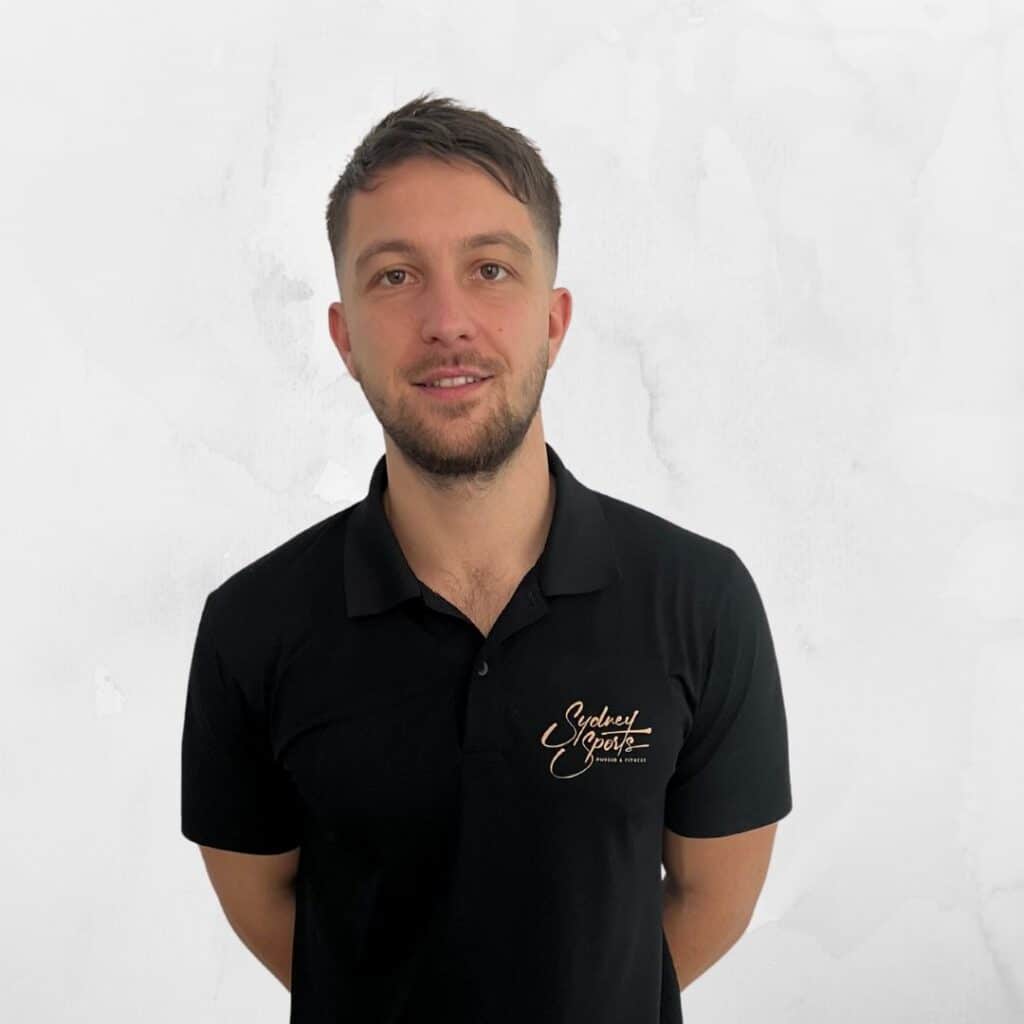 A person wearing a black polo shirt with "Sydney Sports Recovery & Performance" logo stands against a plain white background, smiling slightly, with hands behind their back.