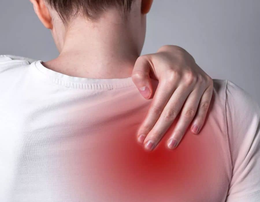 A person is touching their shoulder with one hand, while wearing a white shirt. The shoulder area is highlighted in red, suggesting pain or discomfort. The background is plain gray.