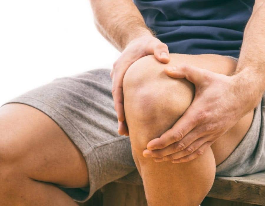 A person sits wearing grey shorts, holding their knee with both hands, suggesting possible discomfort or pain, against a plain background.