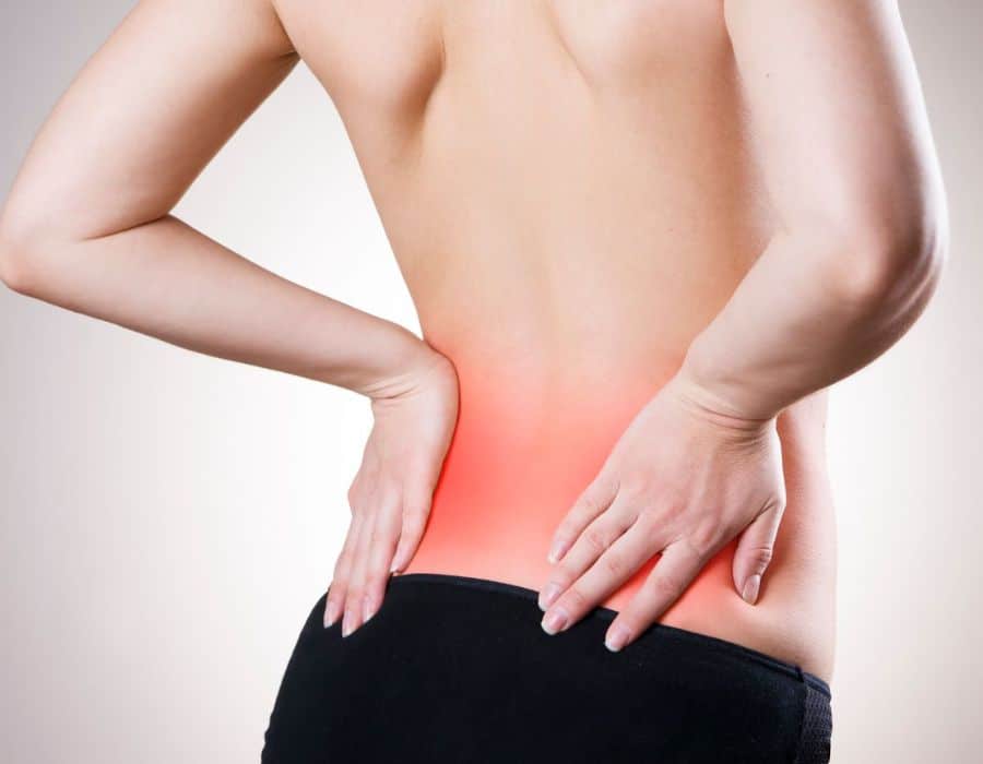 A person holds their lower back, which is highlighted with a red area indicating pain, against a neutral grey background.