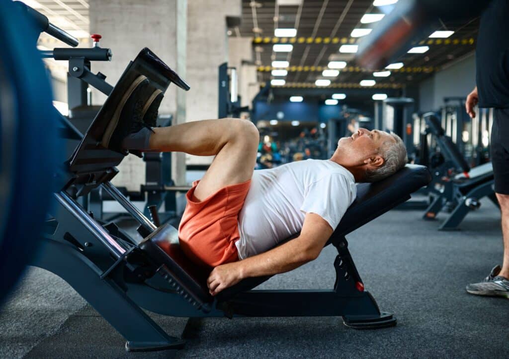 A person in a white shirt and red shorts uses a leg press machine in a gym, surrounded by various exercise equipment and other individuals working out.