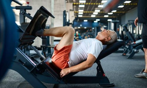 A person in a white shirt and red shorts uses a leg press machine in a gym, surrounded by various exercise equipment and other individuals working out.